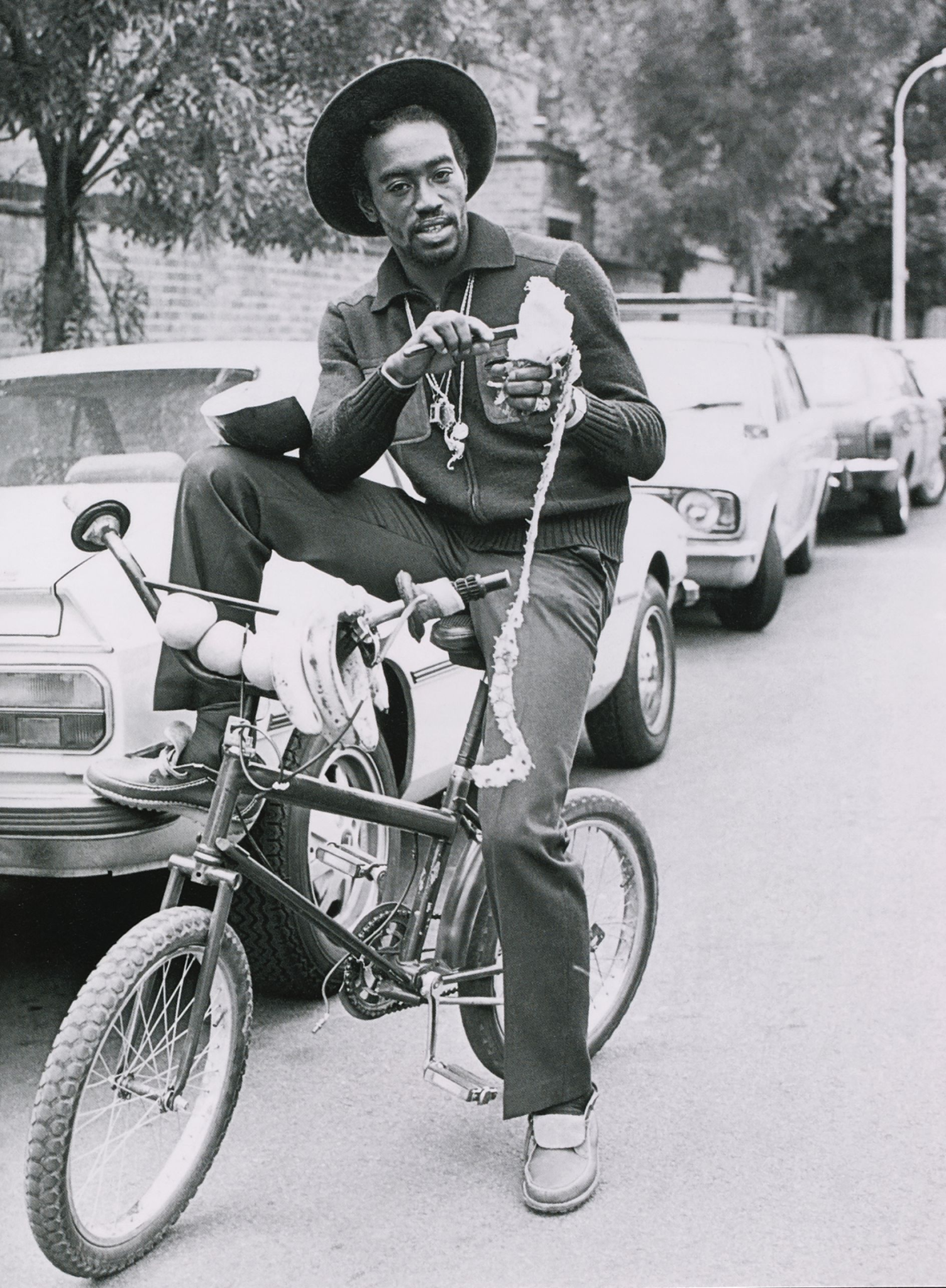 A black-and-white photograph of Deejay Ranking Dread in a wide-brimmed hat, seated on a customised chopper bicycle on a parked-up London street, wearing Clarkes Polyveldt shoes.