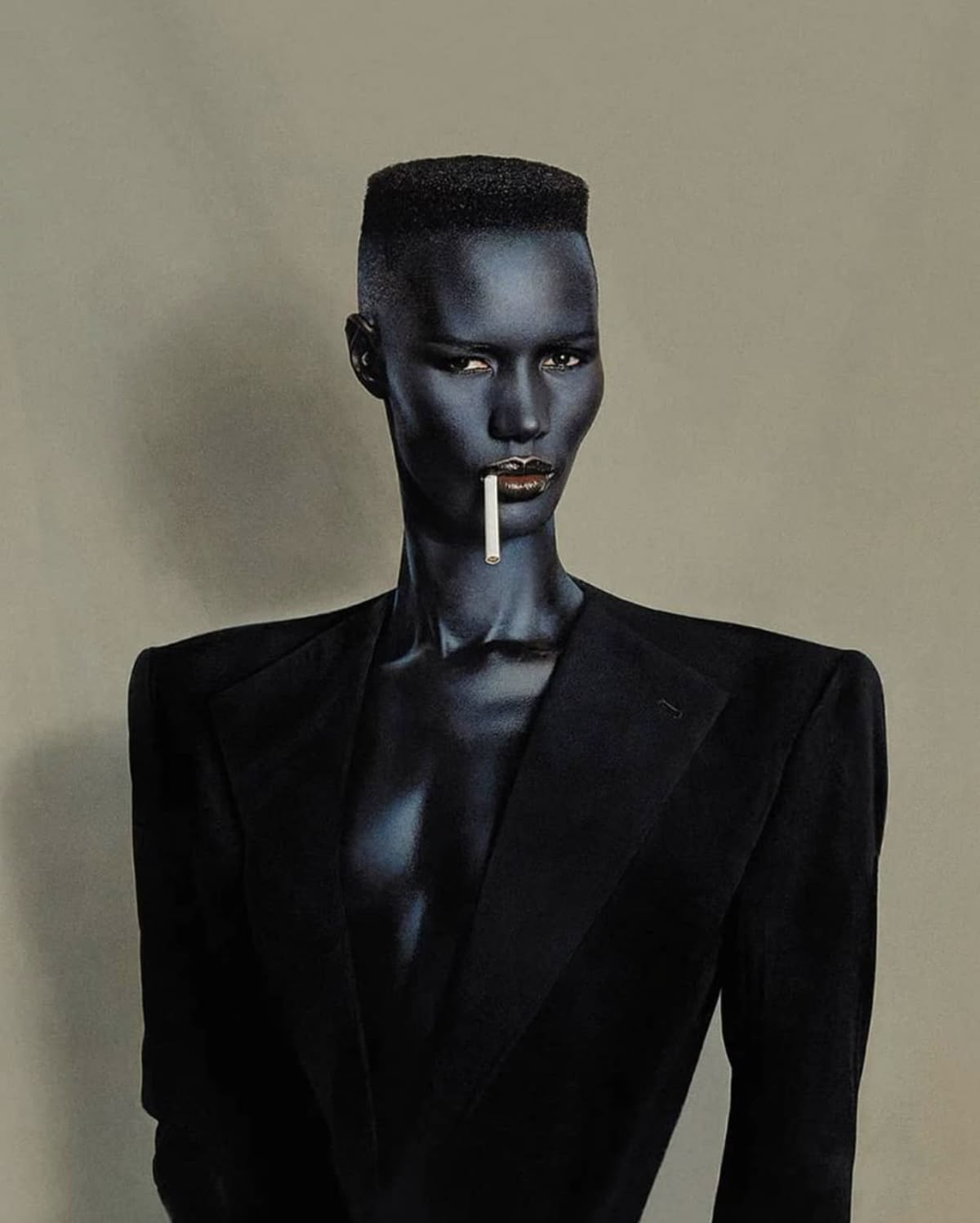 Grace Jones in a black tailored suit with structured shoulders, a cigarette held between her lips, photographed against a plain grey ground.