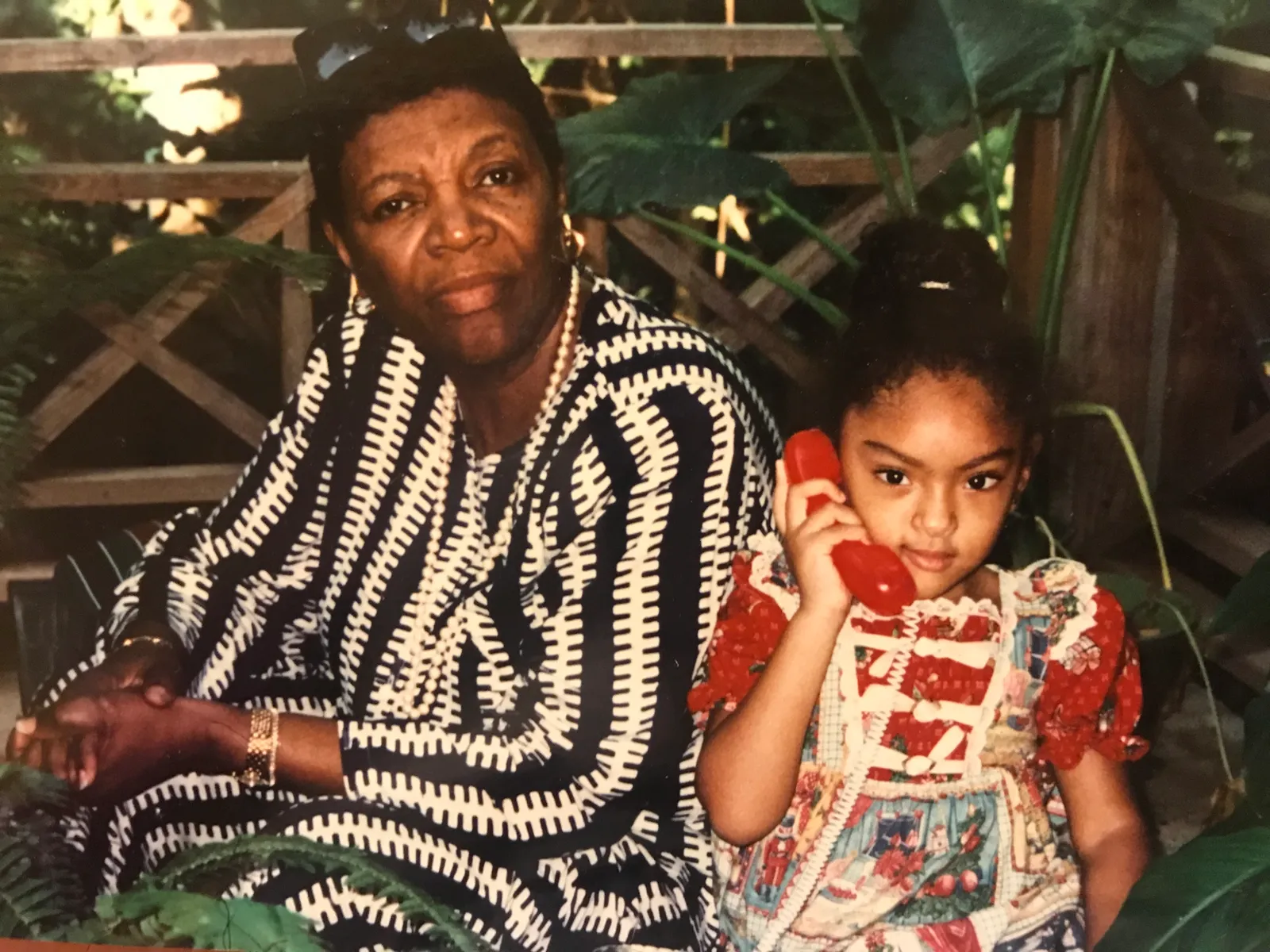 A colour photograph of an older woman in a black-and-white patterned dress seated beside a young girl holding a red telephone, in a tropical garden.