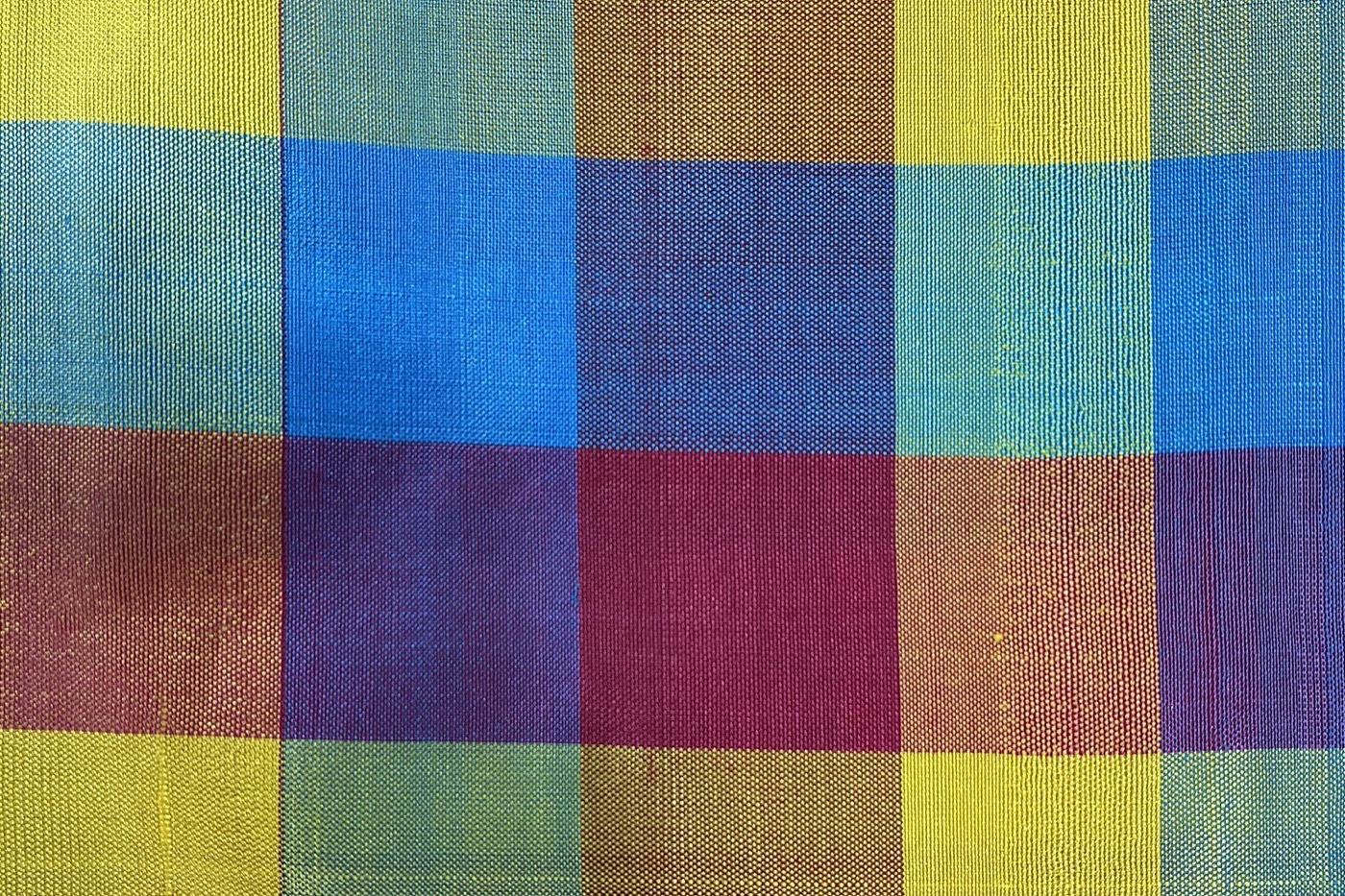 Macro photograph of woven madras check: bands of yellow, blue and red crossing at right angles, with the individual cotton threads visible and the overlaps reading as muddied purple and brown.