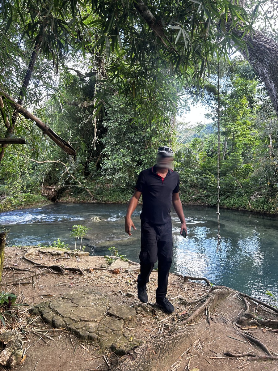 A man in a black polo and cap stands on the rock above the river, phone in hand, the rope hanging beside him