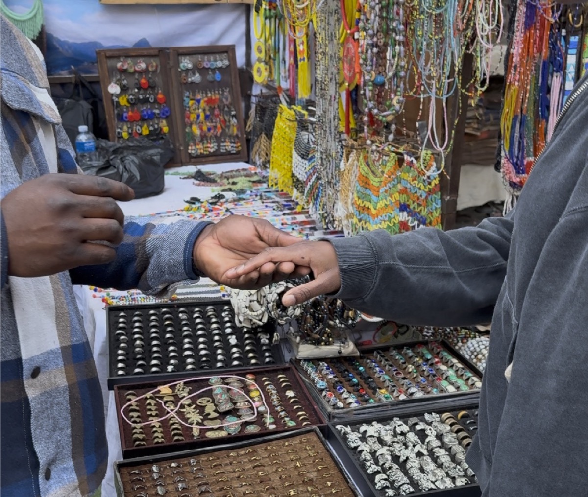 Two hands meeting over a tray of rings at a market stall, the seller's beadwork hung behind