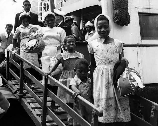 A black-and-white photograph of women and children walking down a ship&rsquo;s gangway, dressed in printed cotton dresses, headscarves and hats, carrying woven baskets and holdalls.